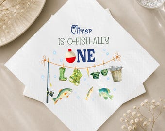 May include: A white paper napkin featuring the text Oliver IS O-FISH-ALLY ONE with a fishing-themed illustration. The design includes a fishing rod, a bobber, a life vest, boots, a bucket of fish, and various colorful fish swimming in blue bubbles.