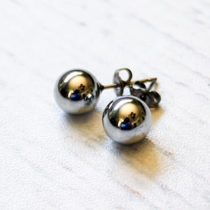 May include: A pair of shiny, silver-colored ball stud earrings. The earrings have a reflective surface, and the posts and backs are visible. The earrings are set against a white and gray background.