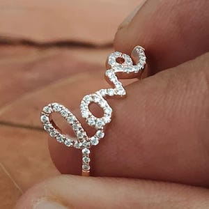 May include: A silver ring spelling out the word "love" in a cursive font. The ring is encrusted with small, clear gemstones. The ring is held between two fingers against a blurred background.