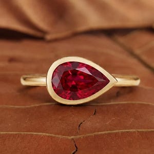 May include: A gold ring with a striking, pear-cut, deep red gemstone. The gem is set in a bezel setting. The ring is presented on a brown, textured surface, highlighting the contrast between the bright gem and the background.