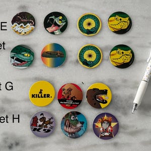 May include: A collection of circular pinback buttons with various cartoon animal designs, including snakes, dogs, and a scorpion. Some buttons have text, such as "KILLER." and "K9". A white pen with a dotted pattern is also present.