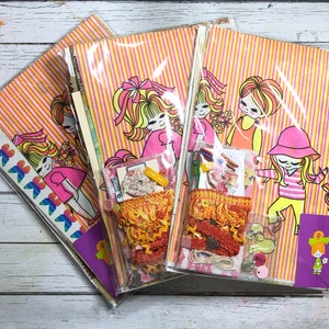 May include: Three sets of colorful patterned paper with illustrations of girls with long hair and bright clothing. Each set includes a variety of embellishments, including ribbons, lace, and buttons.