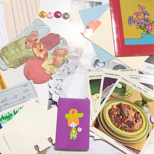 May include: A collection of vintage ephemera, including paper scraps, cards, and a small purple notebook with a girl illustration. The items are arranged on a wooden surface.
