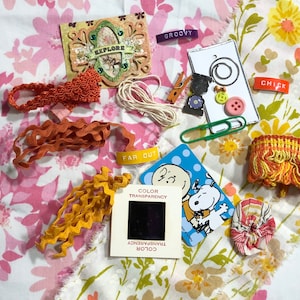 May include: A collection of craft supplies including fabric scraps, ribbon, trim, buttons, and a vintage slide. The fabric scraps feature floral patterns in pink, yellow, and orange. The ribbon is orange and yellow. The trim is orange and yellow. The buttons are red, yellow, and blue. The slide is black and white and features a cartoon image of Snoopy. The text "FAR OUT" and "COLOR TRANSPARENCY" are visible on the slide.
