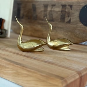 May include: Two brass swan figurines with long necks and curved bodies are displayed on a wooden cutting board. The swans are positioned side-by-side, showcasing their elegant design. The background includes a wooden sign with the word "LAKE".