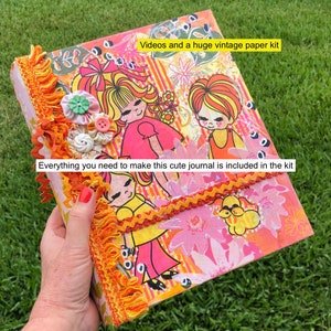 May include: A pink and orange journal with a vintage paper kit design. The cover features illustrations of girls and flowers. The text "Videos and a huge vintage paper kit" and "Everything you need to make this cute journal is included in the kit" is printed on the cover.