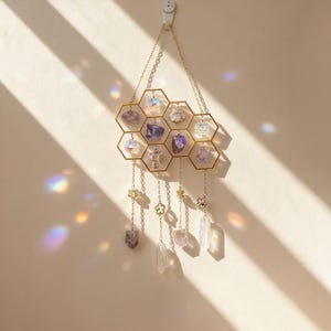 May include: A gold-coloured metal wall hanging with a honeycomb design, holding various crystals and gemstones. The crystals are in shades of purple, clear, and iridescent. Decorative chains with charms hang below. The wall hanging is suspended from a chain.