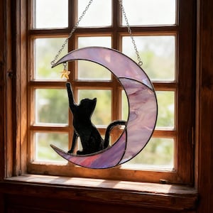 May include: Stained glass crescent moon with a black cat silhouette reaching for a gold star. The moon is a blend of purple and pink hues, hanging from a silver chain. The artwork is displayed in front of a wooden window.
