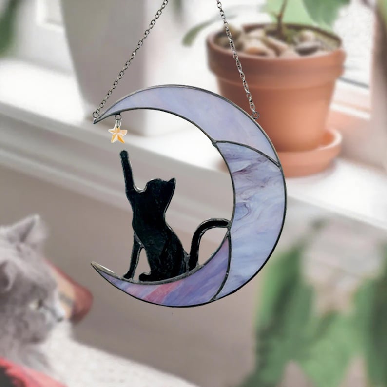 May include: Stained glass crescent moon with a black cat silhouette reaching for a star. The moon features shades of purple and white. A decorative piece for windows or walls.
