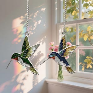 May include: Two stained glass hummingbird ornaments suspended from silver chains. One is green, the other blue and gold. Both have long beaks and hang in front of a window with flowers, catching the light.