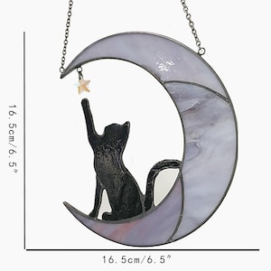 May include: A stained glass cat and moon hanging decoration. The black cat sits on a crescent moon, reaching for a star. The moon is 6.5 inches wide and 6.5 inches tall, with a lavender and white marbled design. A chain is attached for hanging.