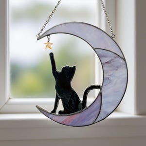May include: A stained glass suncatcher featuring a black cat silhouette reaching for a star. The cat sits on a crescent moon with purple and white hues, suspended by a chain. A charming celestial-themed decoration.