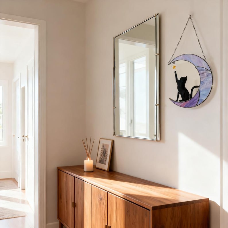 May include: A decorative wall hanging featuring a black cat silhouette reaching for a gold star on a crescent moon. The moon has a stained glass effect with shades of purple and blue. A rectangular mirror and wooden cabinet are also visible.