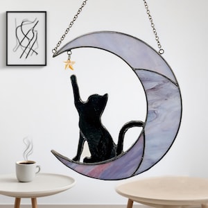 May include: A stained glass cat silhouette reaching for a star, perched on a crescent moon. The moon is a mix of lavender and white, with a chain for hanging. A whimsical decorative piece.