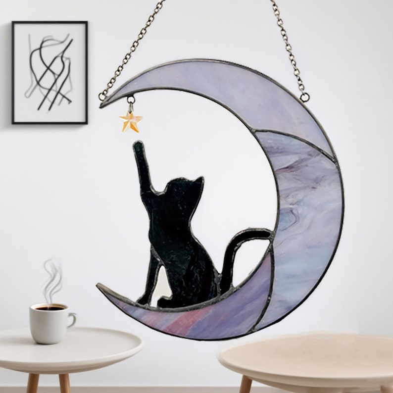 May include: A stained glass cat silhouette reaching for a star, perched on a crescent moon. The moon is a mix of lavender and white, with a chain for hanging. A whimsical decorative piece.
