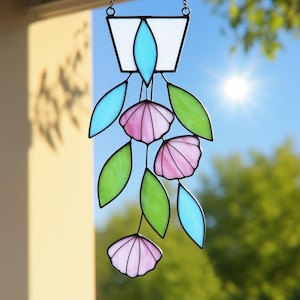 Puede incluir: Un atrapasoles de vidriera con un diseño floral. La pieza presenta una sección superior blanca con forma de lágrima turquesa, flores rosas en forma de concha y hojas verdes y turquesas. El atrapasoles cuelga de una cadena.