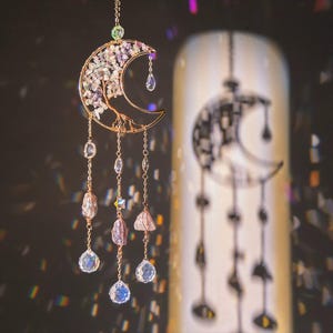 Pode incluir: Um ornamento suspenso com tema celestial, apresentando um design de lua crescente. A lua de metal dourado é adornada com cristais e uma estrutura semelhante a uma árvore. Cristais transparentes e roxos pendem da lua, criando um efeito brilhante. O ornamento é suspenso por uma corrente delicada.