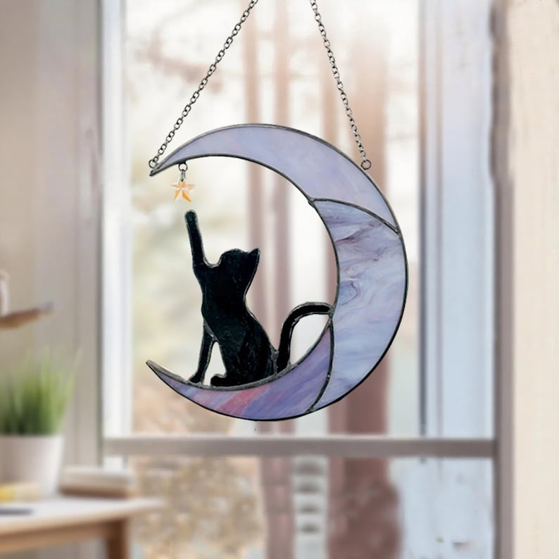 May include: Stained glass crescent moon with a black cat silhouette reaching for a star. The moon is a mix of lavender and white glass, suspended by a chain. A charming cat-themed home decor item.