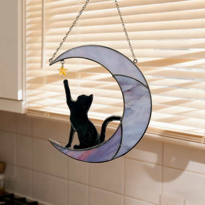 May include: A stained glass crescent moon with a black cat silhouette reaching for a yellow star. The moon has a lavender and purple marbled design, and is suspended by a chain. A whimsical cat-themed decoration.