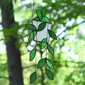 May include: A stained glass hanging planter with a white pot and vibrant green leaves. The leaves are a mix of solid and textured glass, with black outlines. The planter hangs from a delicate chain, creating a charming, nature-inspired decoration.