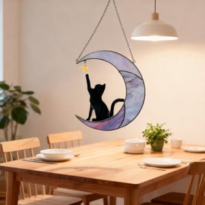May include: A stained glass crescent moon with a black cat silhouette reaching for a yellow star. The moon has purple and white hues and hangs from a silver chain. A decorative piece for a window or wall.