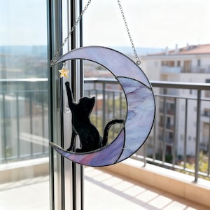 May include: A stained glass crescent moon with a black cat silhouette reaching for a gold star. The moon has a purple and white marbled design and is suspended by a silver chain. A decorative piece for cat lovers.