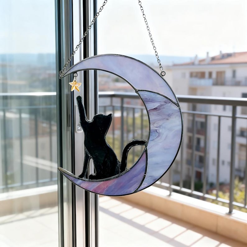 May include: A stained glass crescent moon with a black cat silhouette reaching for a gold star. The moon has a purple and white marbled design and is suspended by a silver chain. A decorative piece for cat lovers.