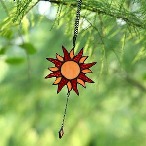 May include: A stained glass suncatcher with a sun design. The sun has a central orange circle, surrounded by red and orange rays. It hangs from a black chain and is suspended against a blurred green background.