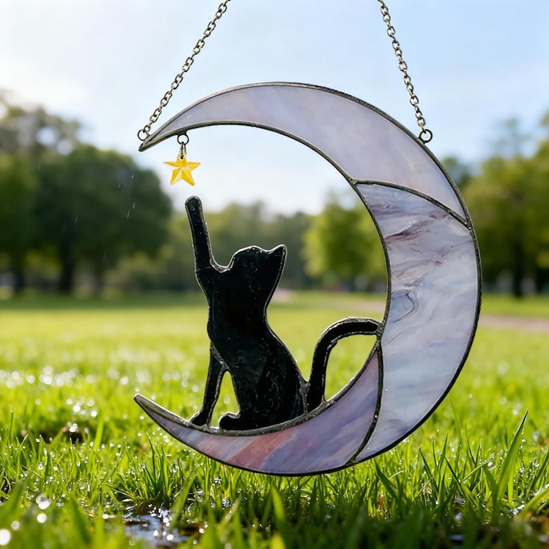 May include: A stained glass crescent moon with a black cat silhouette reaching for a yellow star. The moon has a white and purple marbled design and hangs from a chain. The cat is sitting on the moon.