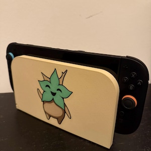 May include: A black Nintendo Switch console is docked in a beige stand. The stand features a cartoon image of a green and brown plant-like character. The stand is made of a light beige material.