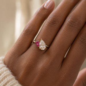 Può includere: Un anello con una pietra preziosa trasparente a taglio pera e una pietra rosa più piccola a taglio pera su una sottile fascia metallica. Le due pietre sono posizionate fianco a fianco in un'incastonatura asimmetrica, creando un netto contrasto.