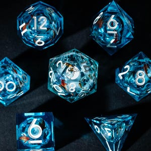 May include: A set of seven translucent blue polyhedral dice with white numbering. The dice have a shimmering, aquatic appearance, with small, colorful inclusions. The dice are arranged on a dark surface, with light reflecting off their facets.