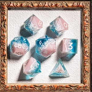 Puede incluir: Juego de siete dados poliédricos en un marco decorativo. Los dados son una mezcla de rosa, azul y blanco, con números visibles. Los dados están diseñados para juegos de mesa. El marco es marrón con detalles ornamentados.