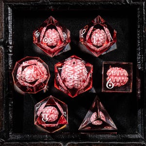 May include: A set of seven polyhedral dice made of transparent red resin, each containing a miniature white brain model inside. The dice feature white numbers and symbols, including a star icon on the d6 and d100, arranged in a display.