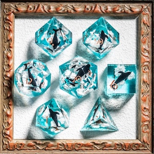 May include: A set of seven polyhedral dice with a marine theme, displayed in a decorative wooden frame. Each die is transparent blue with a black dolphin design and white cloud accents. The numbers are in a copper color.