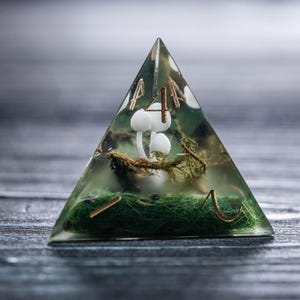 Pode incluir: Um peso de papel triangular transparente de resina com uma cena florestal em miniatura. Cogumelos brancos, musgo verde e detalhes dourados est&atilde;o encapsulados dentro da pir&acirc;mide transparente. O topo apresenta letras douradas. A est&eacute;tica geral &eacute; caprichosa e inspirada na natureza.