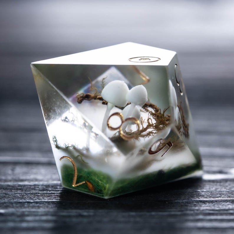 Pode incluir: Um dado de resina transparente de v&aacute;rias faces com figuras de cogumelos brancos, detalhes dourados e musgo verde. O dado tem n&uacute;meros dourados e est&aacute; sobre um fundo escuro. Um acess&oacute;rio de jogo de mesa &uacute;nico.