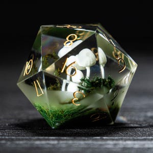 Pode incluir: Um dado transparente multifacetado com numera&ccedil;&atilde;o dourada e uma cena interna. A cena apresenta cogumelos brancos e musgo verde. O dado est&aacute; sobre um fundo escuro, destacando a paisagem em miniatura.