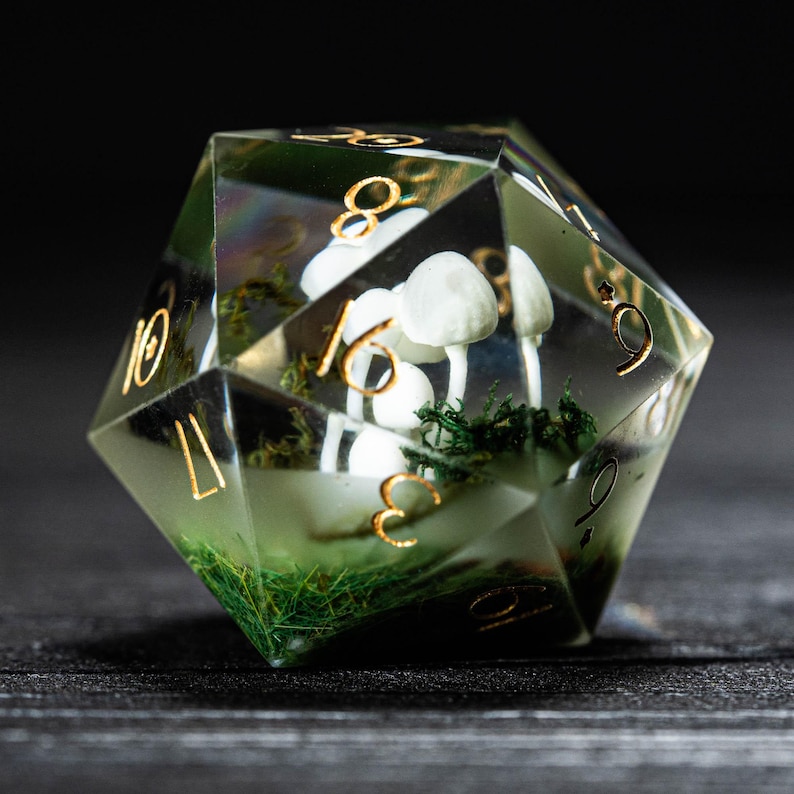 Pode incluir: Um dado transparente multifacetado com numera&ccedil;&atilde;o dourada e uma cena interna. A cena apresenta cogumelos brancos e musgo verde. O dado est&aacute; sobre um fundo escuro, destacando a paisagem em miniatura.