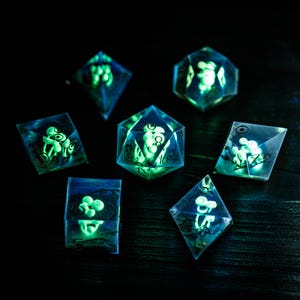 May include: A set of seven glowing dice in various geometric shapes. The dice are transparent with a blue hue and contain a glowing green design. The dice are arranged on a dark surface, creating a striking contrast.