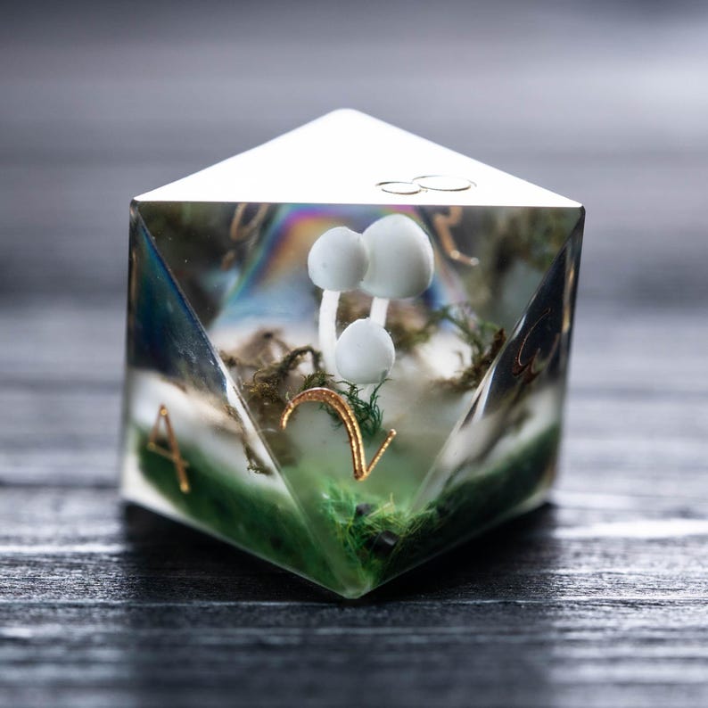 Pode incluir: Um dado de resina transparente e facetado com uma cena de floresta em miniatura no interior. A cena inclui figuras de cogumelos brancos, musgo verde e detalhes dourados. O dado tem n&uacute;meros dourados e est&aacute; sobre um fundo escuro.