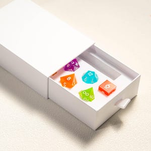 Pode incluir: Uma caixa branca com uma gaveta parcialmente aberta, revelando seis dados poli&eacute;dricos coloridos. Os dados s&atilde;o de v&aacute;rias cores, incluindo roxo, laranja, turquesa, verde e vermelho. Estes dados s&atilde;o usados para jogos de tabuleiro.