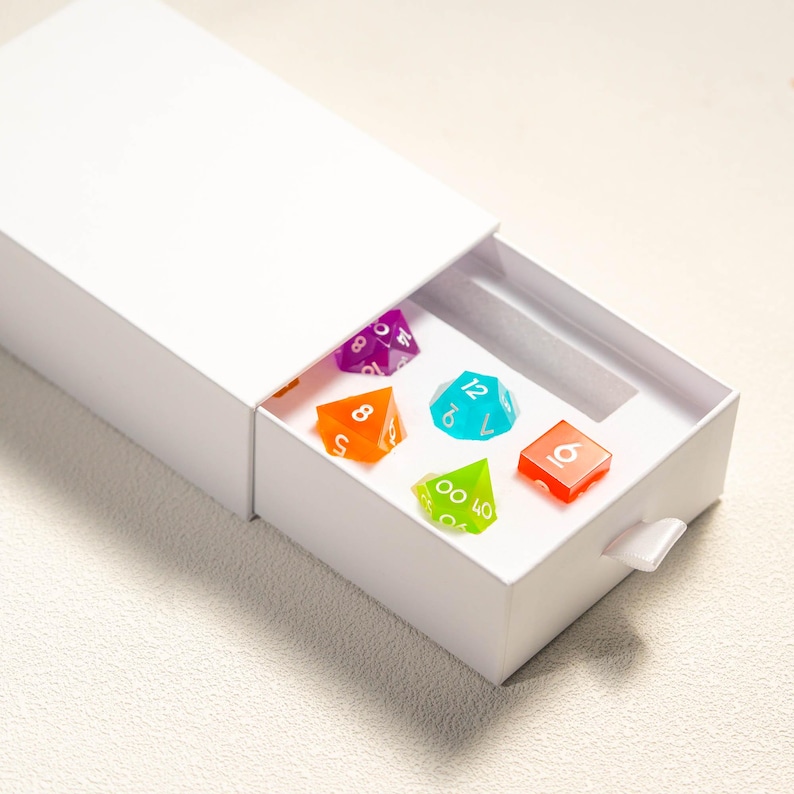 Pode incluir: Uma caixa branca com uma gaveta parcialmente aberta, revelando seis dados poli&eacute;dricos coloridos. Os dados s&atilde;o de v&aacute;rias cores, incluindo roxo, laranja, turquesa, verde e vermelho. Estes dados s&atilde;o usados para jogos de tabuleiro.