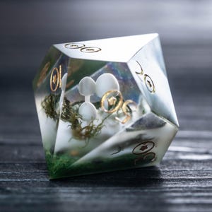 Pode incluir: Um dado D20 transparente e multifacetado com uma cena de floresta em miniatura no interior. A cena inclui figuras de cogumelos brancos, musgo verde e detalhes dourados. O dado tem n&uacute;meros dourados e est&aacute; sobre um fundo escuro.