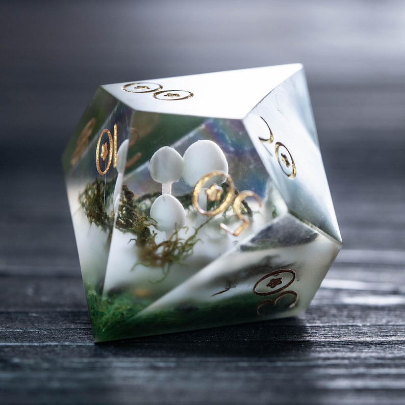 Pode incluir: Um dado D20 transparente e multifacetado com uma cena de floresta em miniatura no interior. A cena inclui figuras de cogumelos brancos, musgo verde e detalhes dourados. O dado tem n&uacute;meros dourados e est&aacute; sobre um fundo escuro.
