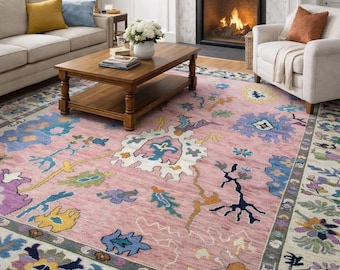 May include: A living room featuring a large, pink area rug with a floral pattern in shades of blue, yellow, and purple. The rug sits beneath a wooden coffee table and in front of a fireplace. A beige sofa and armchair complete the scene.