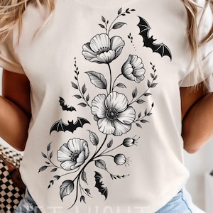 May include: Cream-colored t-shirt featuring a black floral and bat design. The graphic includes stylized flowers and bats, creating a gothic or Halloween theme. The text "THE QUIET NIGHT" is visible at the bottom of the shirt.