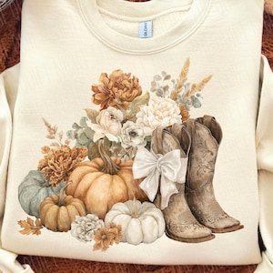 May include: Cream-colored sweatshirt featuring a watercolor design of pumpkins, flowers, and cowboy boots. The design includes a white bow and various fall-themed elements. The long-sleeved top is a cozy fall fashion item.