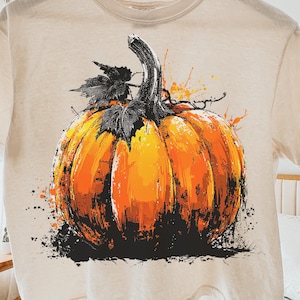May include: A cream-colored t-shirt featuring a vibrant, artistic pumpkin design. The pumpkin is rendered in shades of orange and yellow with black accents, and a dark green stem. The shirt is hanging on a wooden hanger.