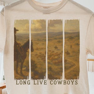 May include: Cream-colored t-shirt featuring a Western-themed graphic. The graphic depicts a cowboy on horseback in a landscape, divided into vertical panels. The text "LONG LIVE COWBOYS" is printed below the image. The shirt is hanging on a wooden hanger.